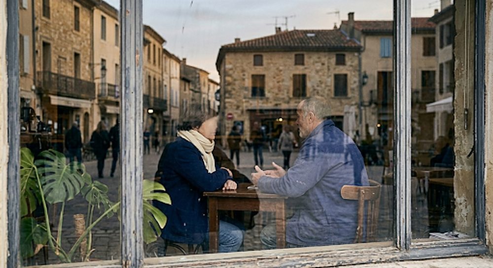 Deux personnes en discussion à une table de café, vues de la rue à travers la vitrine